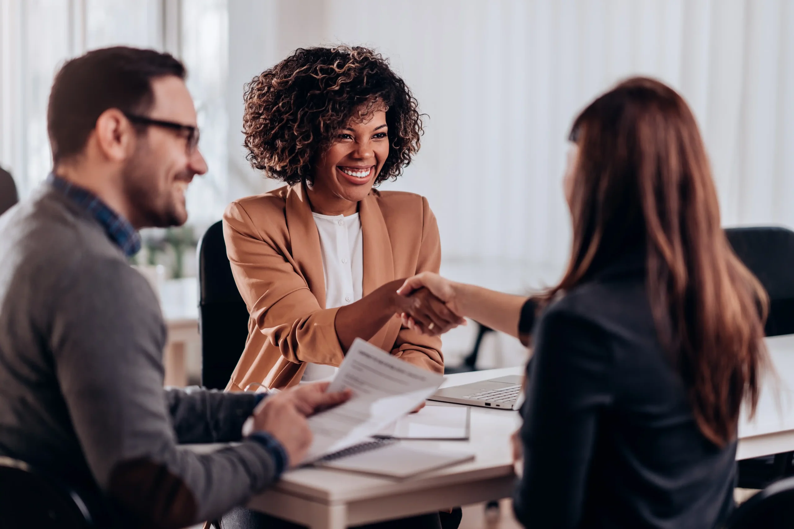 Smiling woman shakes the hand of another woman while a man holding papers smiles at the both of them.