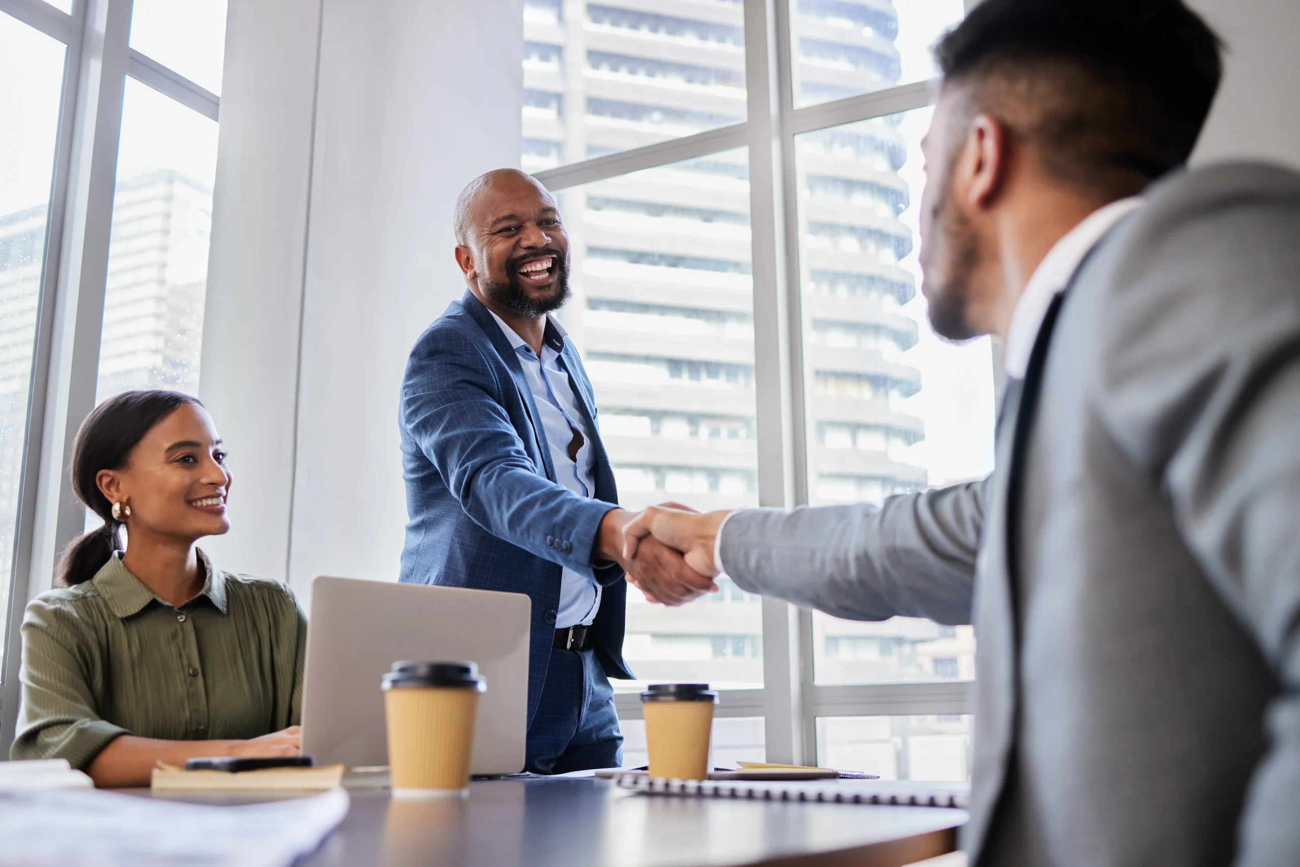 Business man shaking hand with another man across a table while woman sits and smiles at the two men.